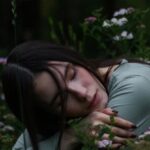 Vertical Gardens - Young female with dark hair embracing herself and putting head on hands sitting on squat in grass with blooming meadow flowers in park