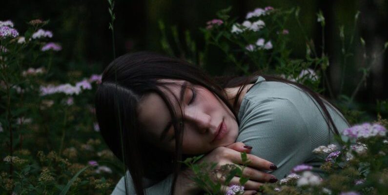 Vertical Gardens - Young female with dark hair embracing herself and putting head on hands sitting on squat in grass with blooming meadow flowers in park