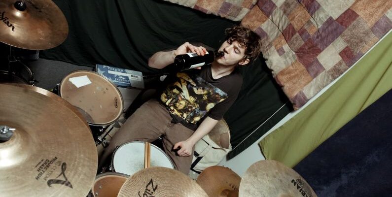 Soundproofing - From above of male musician in casual outfit sitting at drum set near wall with bottle of beer in studio