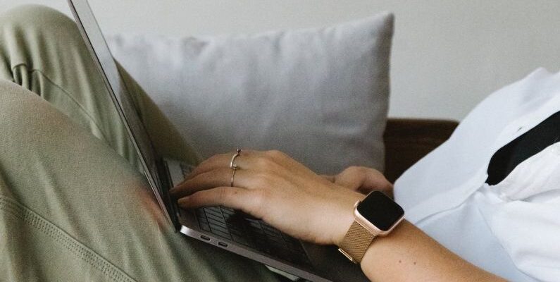 Smart Home Sensors - Side view of crop unrecognizable female freelancer in casual clothes and smartwatch lying on comfortable sofa and working remotely on laptop